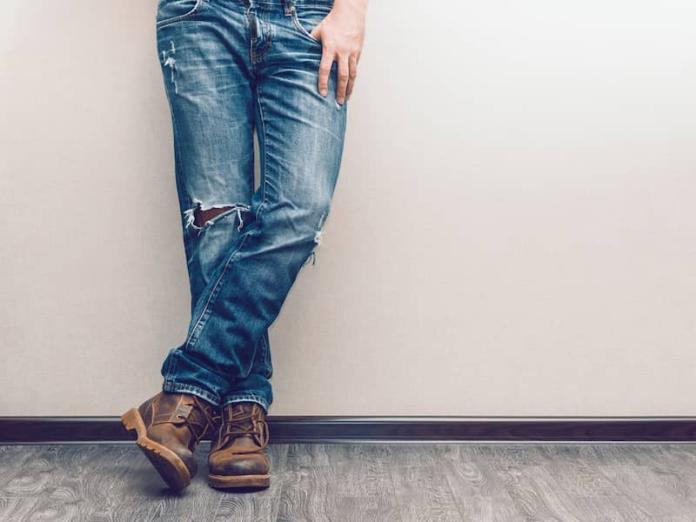 The bottom half of a man wearing faded jeans and leaning against a white wall