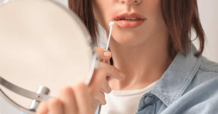 a woman looking into a small mirror and dabbing at a cold sore with a cotton swab