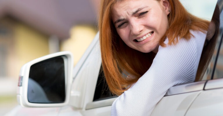 A redheaded woman in a white long sleeved shirt looking backward out of the driver's side window of a white car. She is cringing.