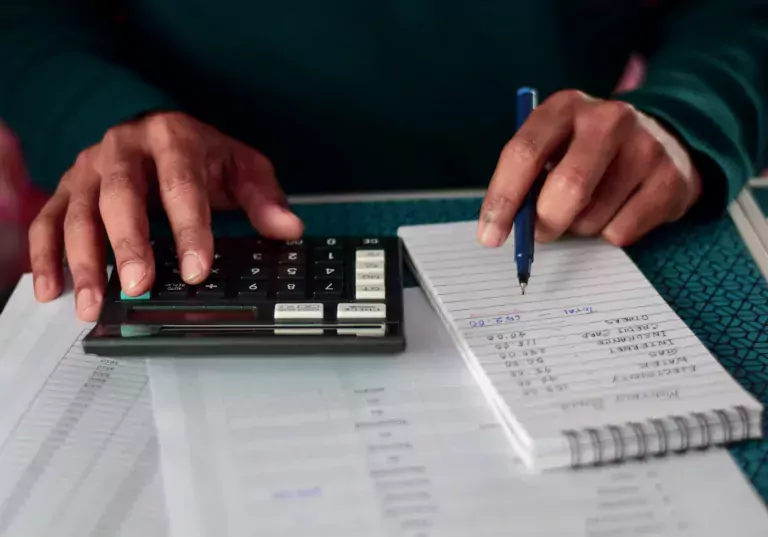Hands with a calculator and a pad of paper with budget items listed.