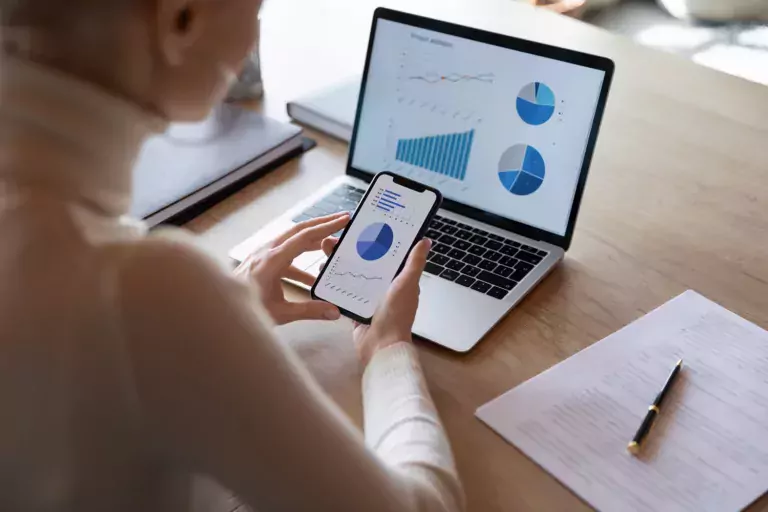 Woman hold smartphone use pc at workplace. Project stats financial data sales charts on laptop and cellphone screen, close up view over shoulder.