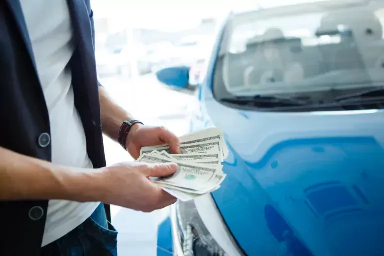 A blue car and someone standing in front of it with dollar bills in hand