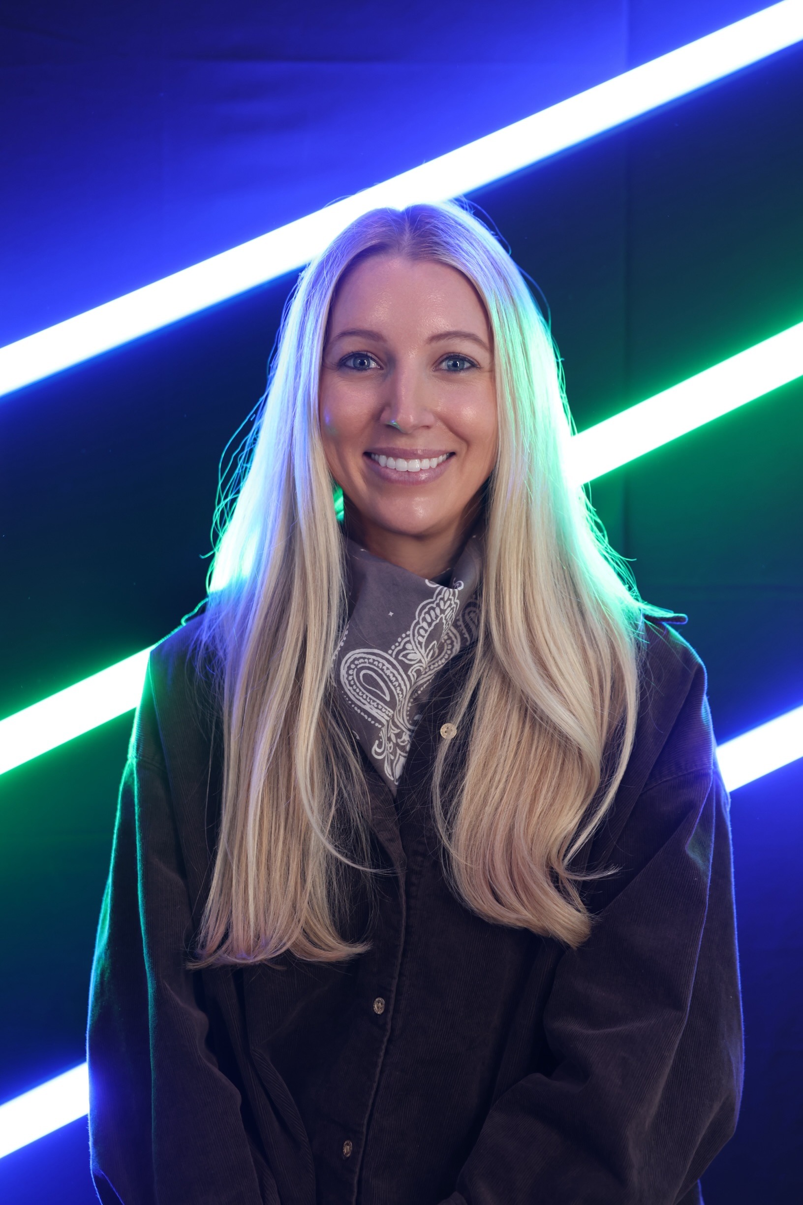 Woman with long blond hair, wearing black jacket. Blue and green neon lights behind.